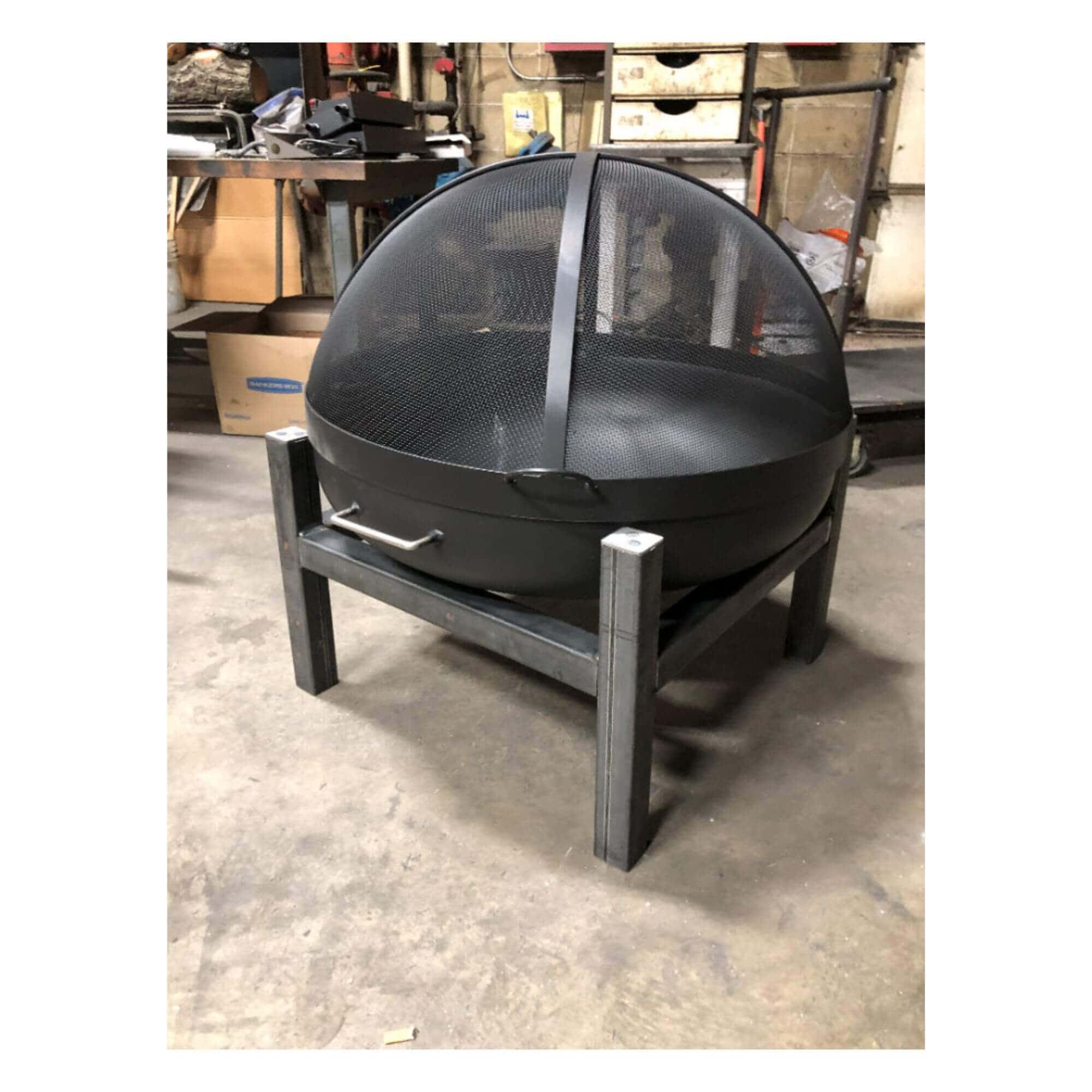 A black metal fire pit with a dome-shaped mesh screen and a square support stand, photographed in a workshop or garage setting.