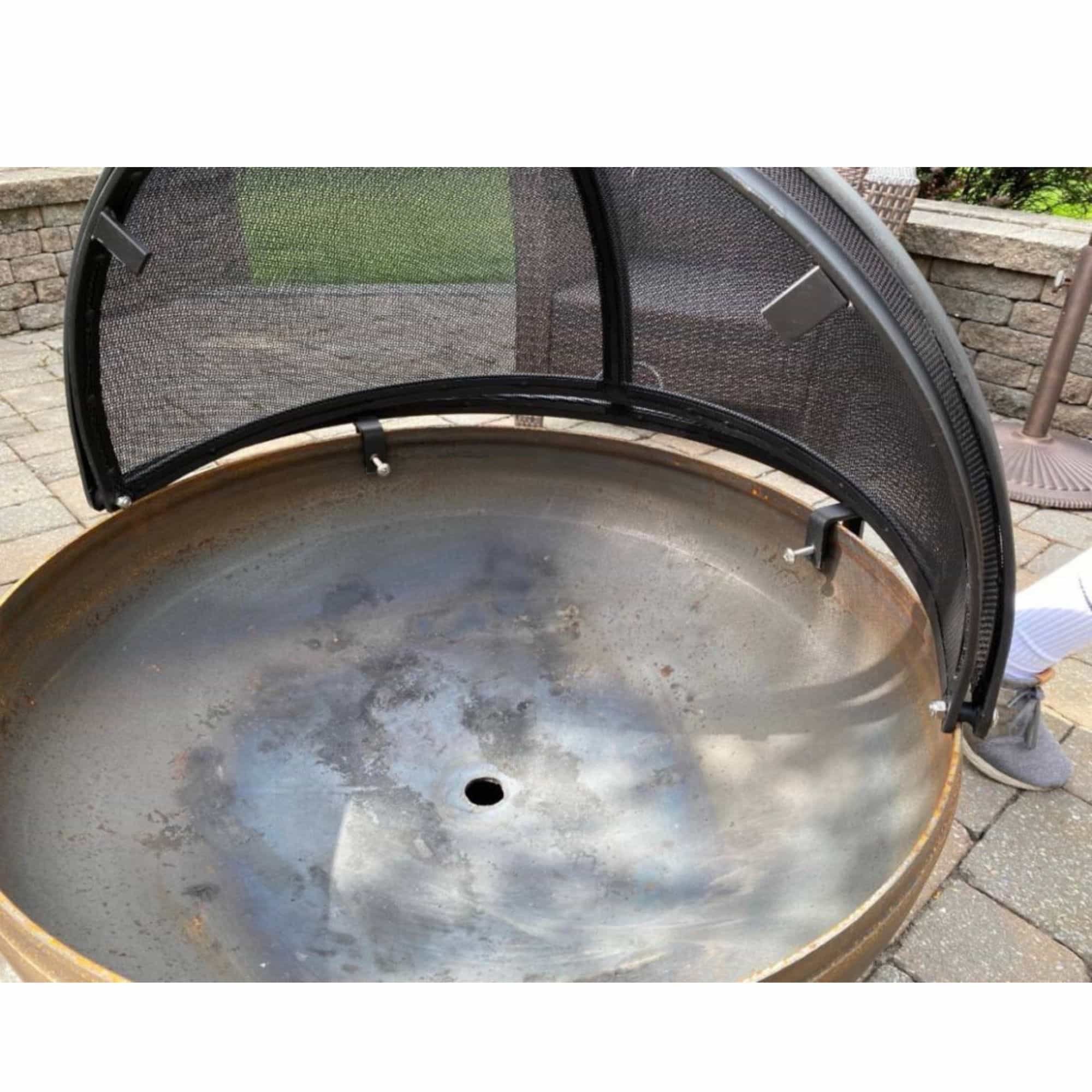 A round metal fire pit or grill base with a black mesh safety screen partially lifted, shown outdoors on a paved patio with a stone wall and greenery in the background.
