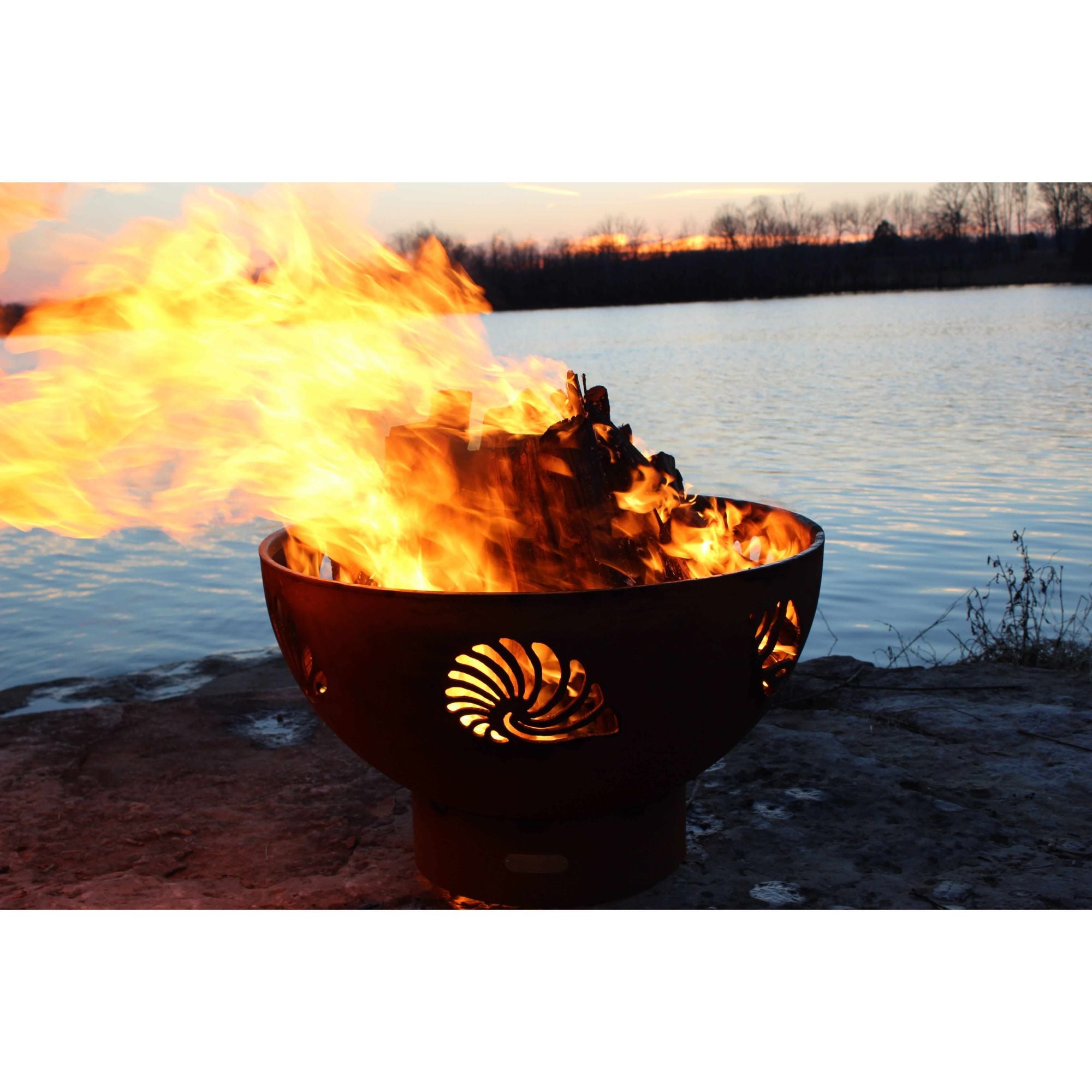 Fire pit with flames burning brightly by a body of water at sunset.