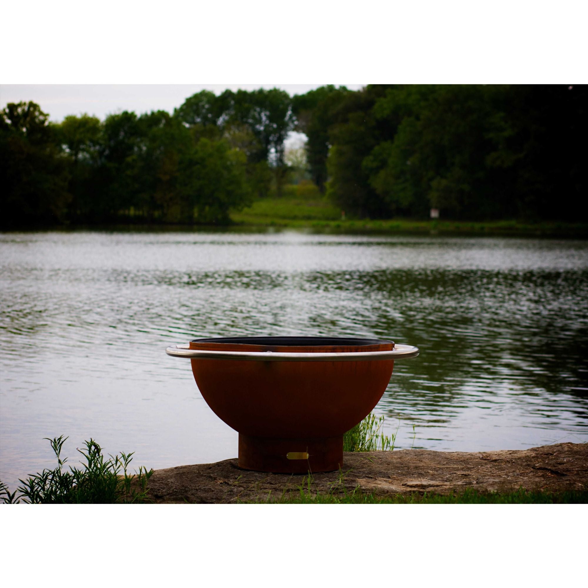 Rust-colored fire pit by a lake with trees in the background