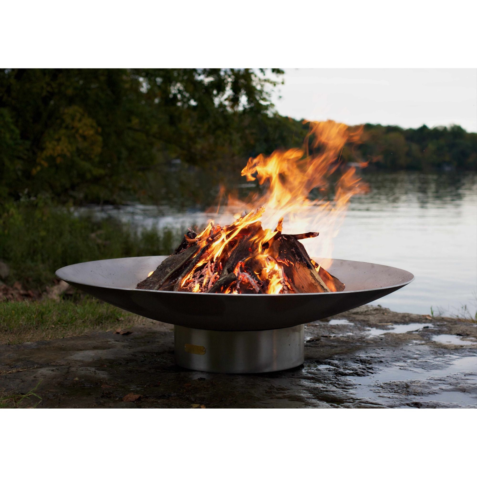 Fire pit with flames burning in a natural setting by a body of water.