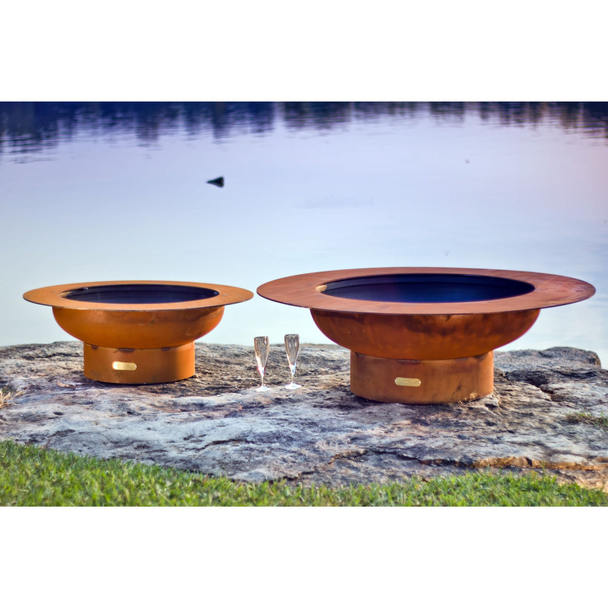 Two metal fire pits on a rocky outcrop by a body of water.