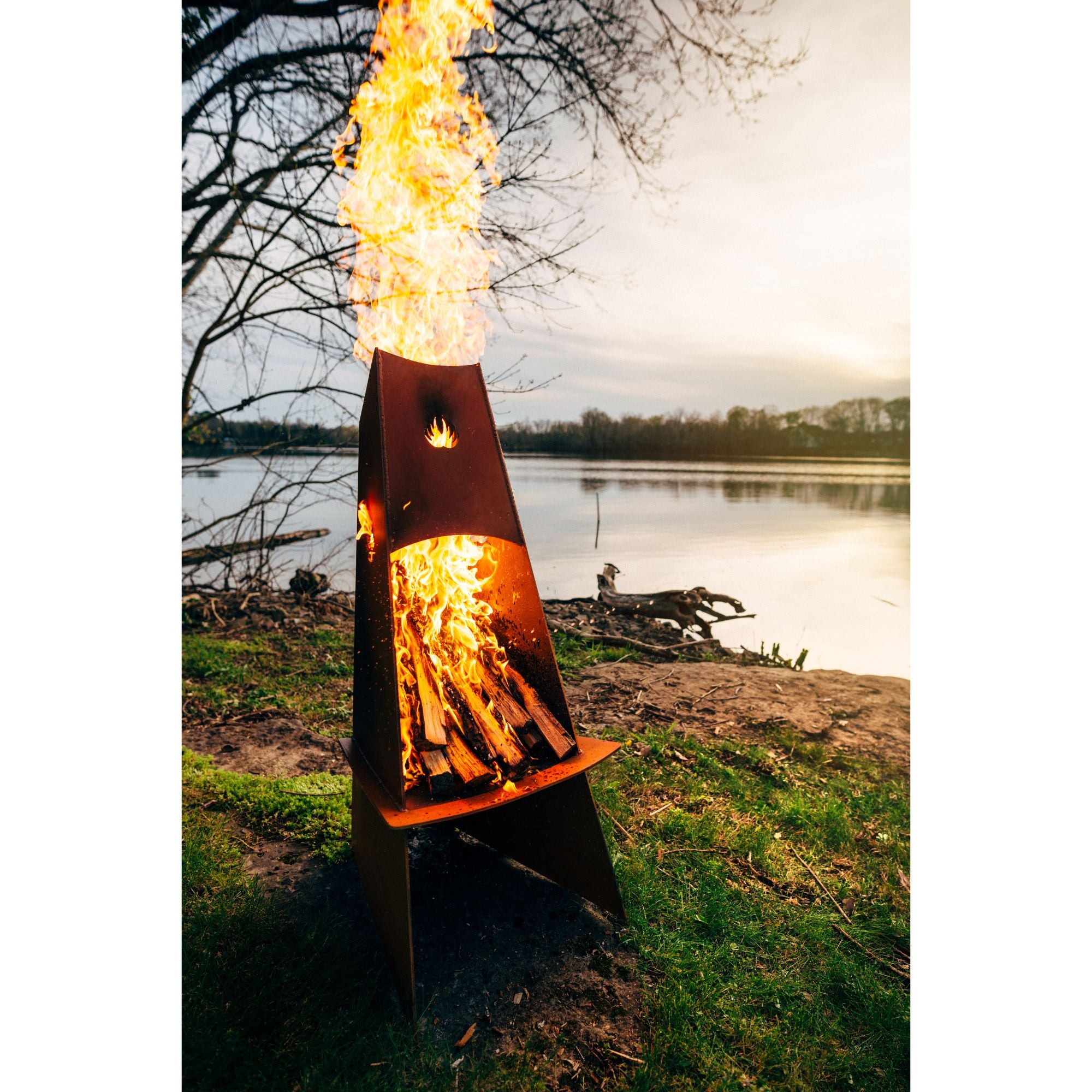 Tall metal fire pit with flames lit outdoors by a lake