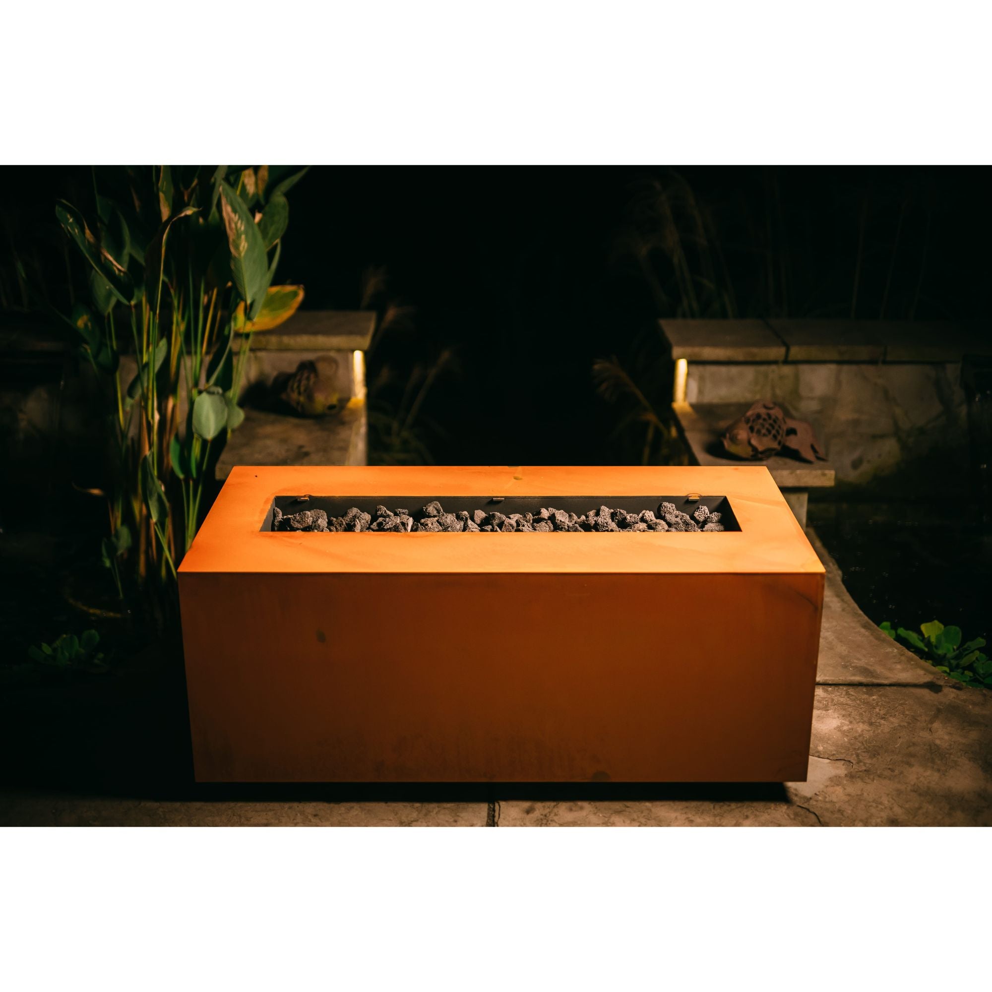 Rectangular fire pit with orange finish in a garden setting at night.
