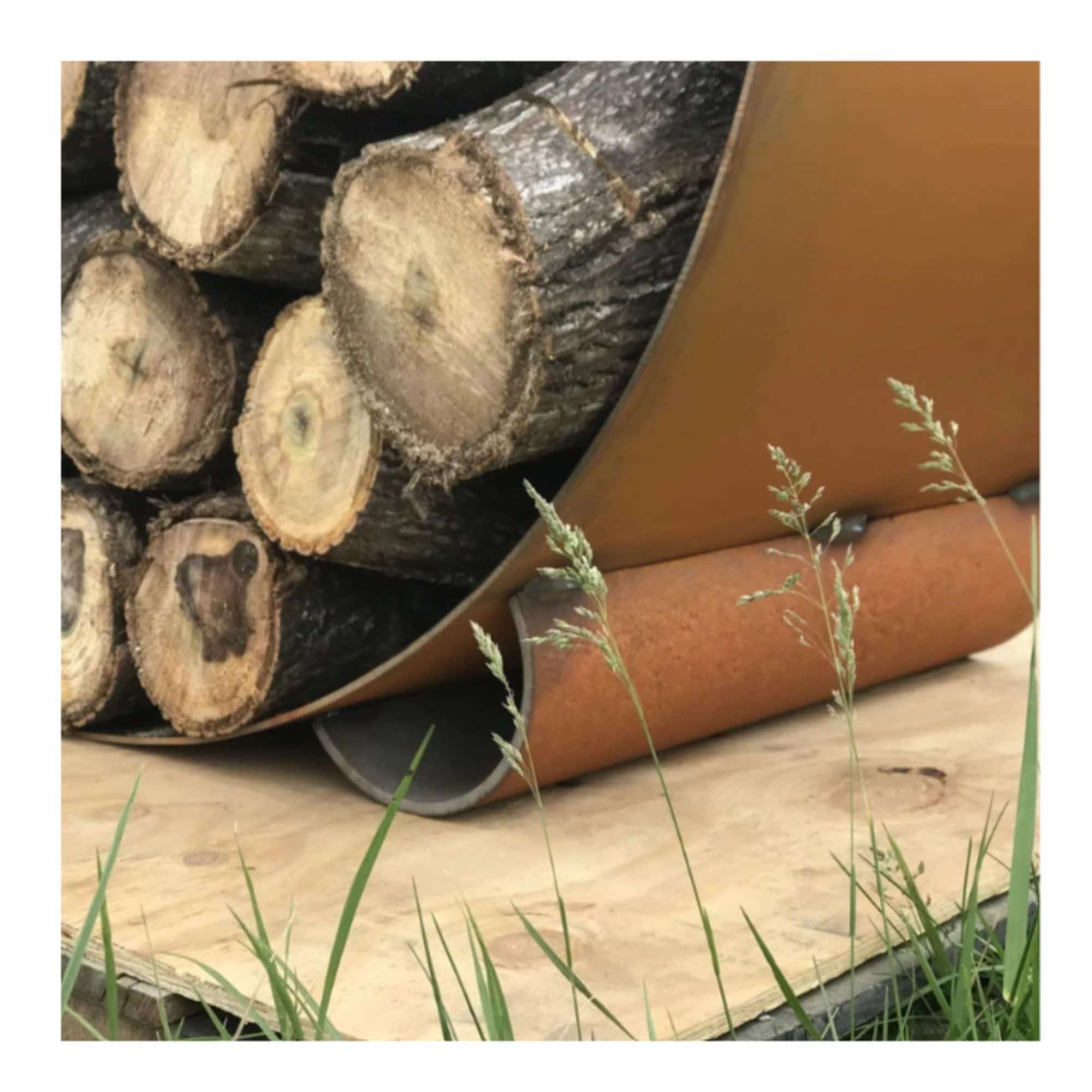 Stack of logs in a steel round log rack on a wooden surface, surrounded by grass.