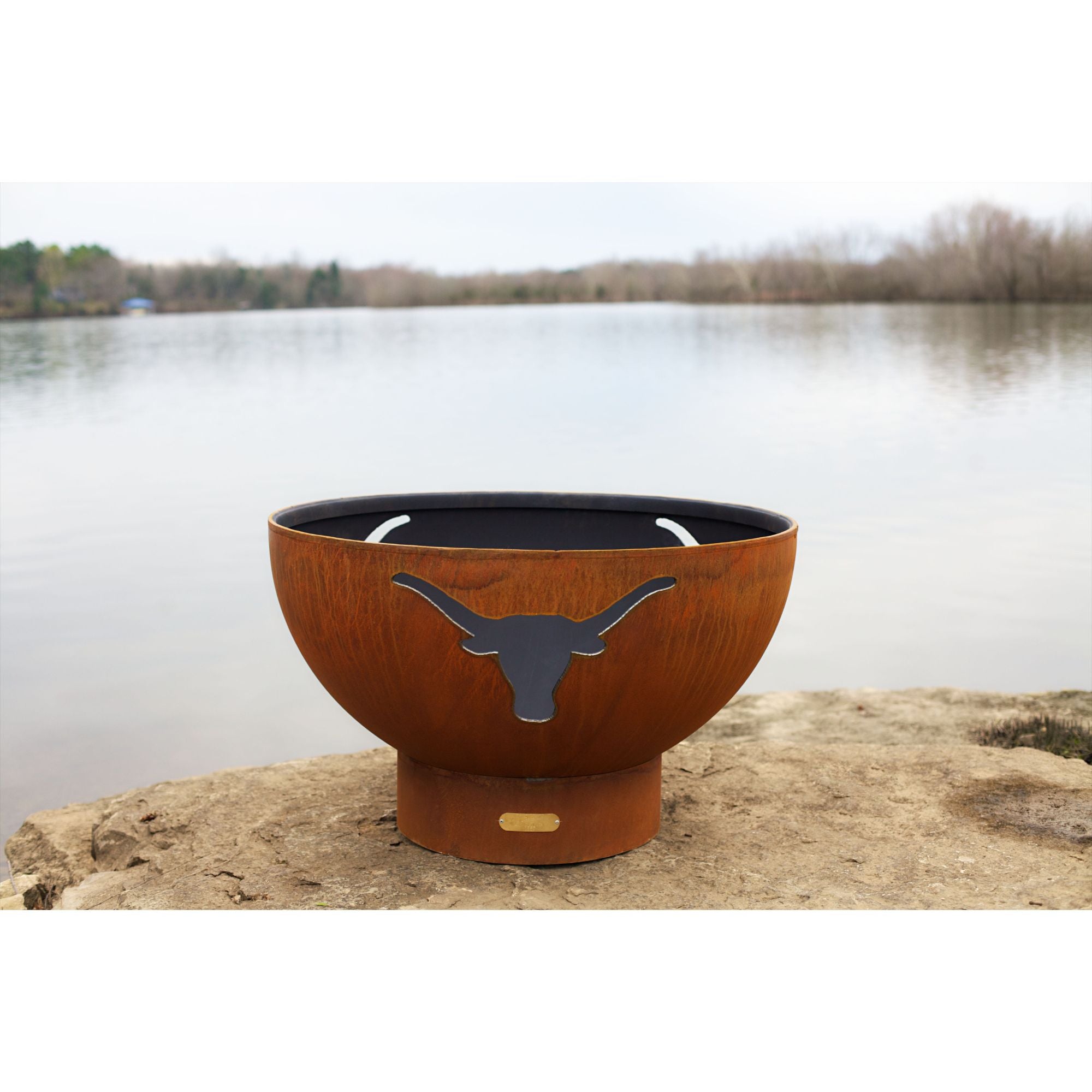 Rust-colored fire pit with longhorn design on a rock by a lake