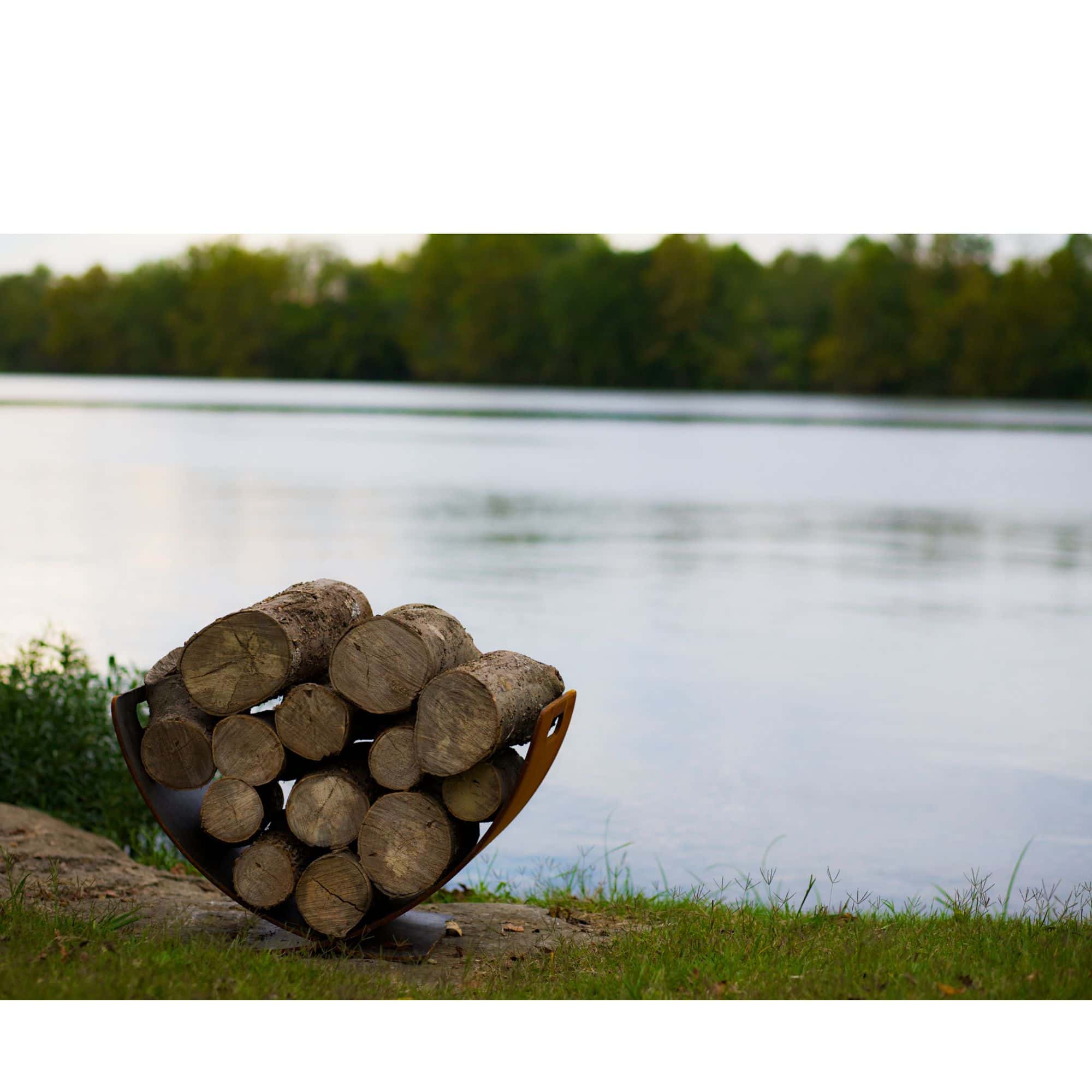 Stack of logs arranged in a circular pattern by a lake with trees in the background