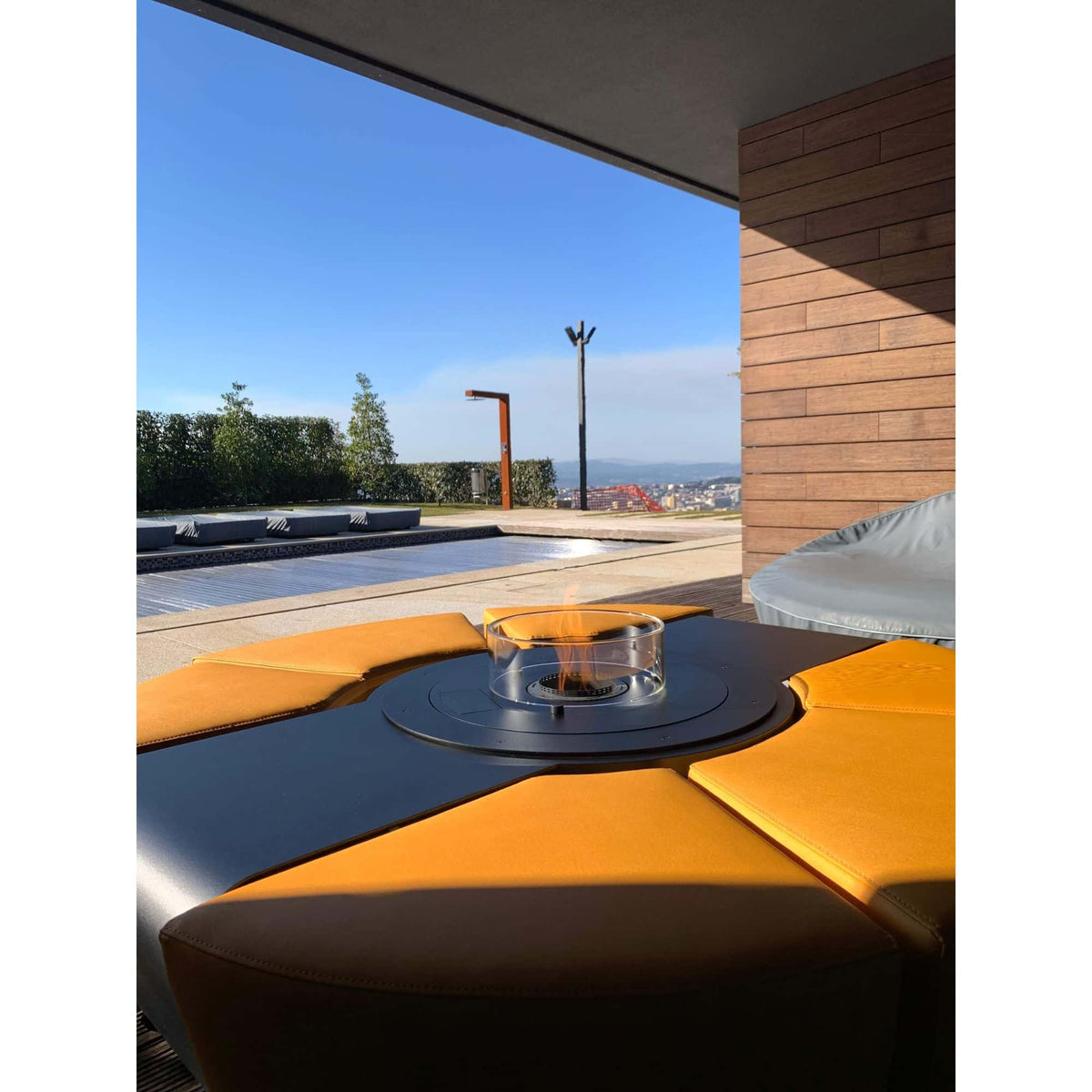 Outdoor patio furniture featuring a round black fire pit table with a glass wind guard and visible flame, surrounded by a modern modular sectional sofa with mustard yellow cushions, set under a covered patio overlooking a pool and city view.