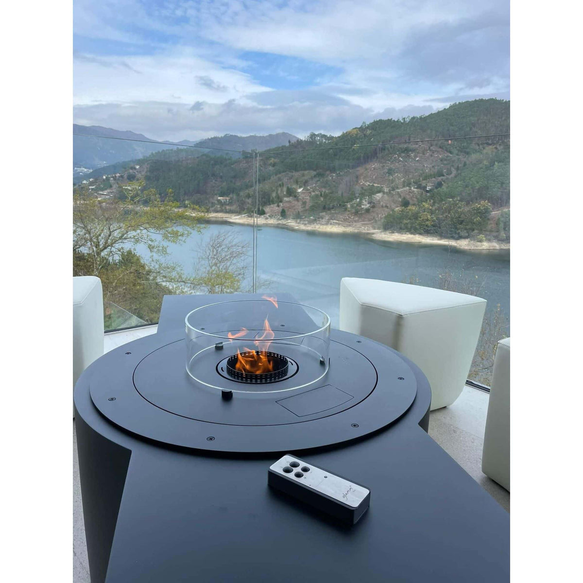 A modern outdoor fire pit table with a clear glass wind guard and active flame, set on a patio with a remote control. The background shows a scenic view of a lake and mountains through a glass railing.