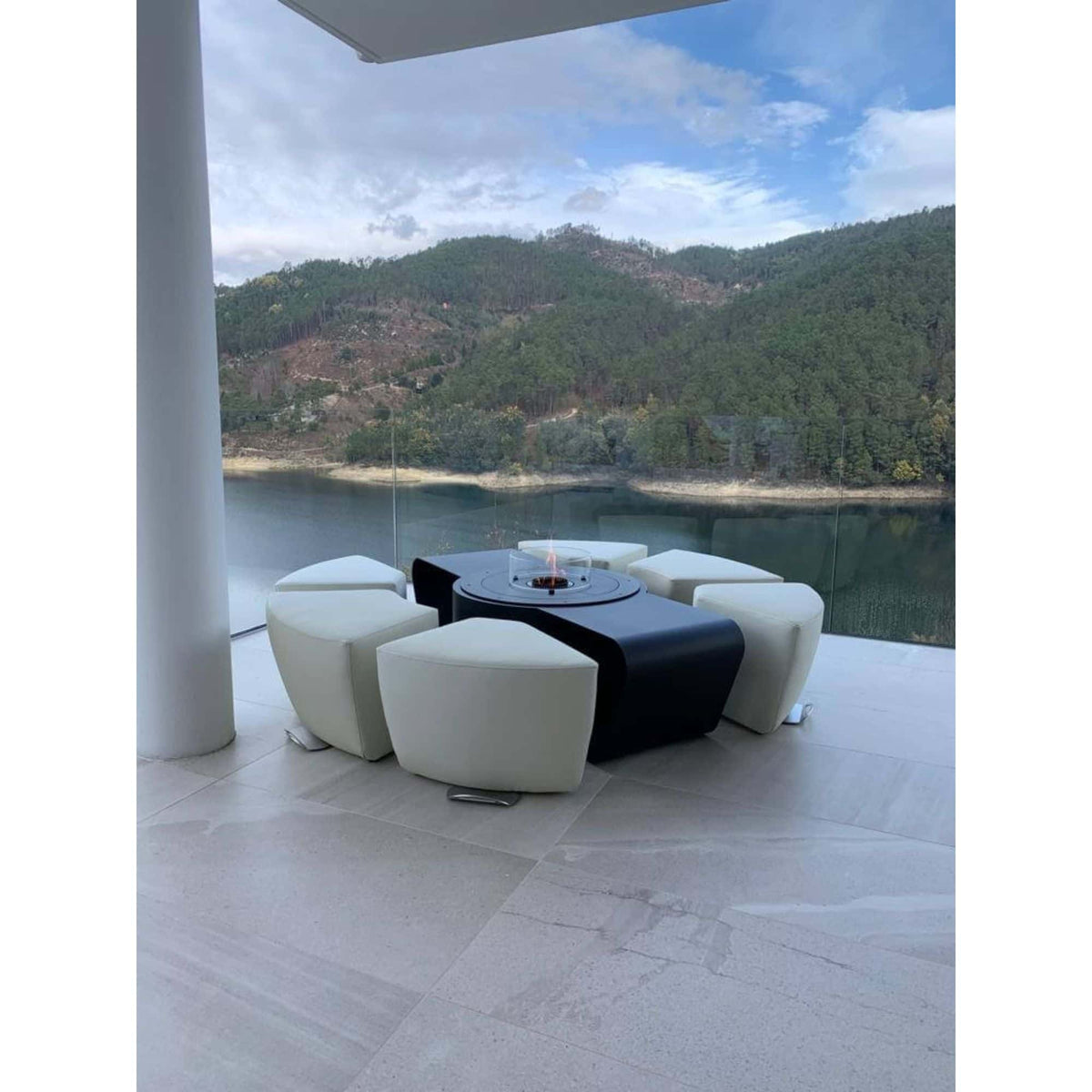 Modern outdoor patio furniture set featuring a round black fire pit table surrounded by six curved white bucket-style stools, set on a light stone tile deck with a scenic mountain and lake view in the background.