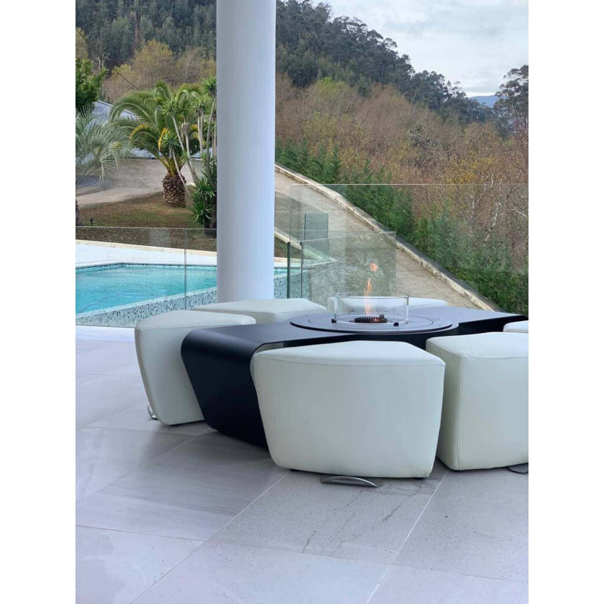 Modern outdoor patio furniture set featuring a black round fire pit table with a clear glass wind guard, surrounded by four chunky white leather-like ottomans, set on a light gray tiled patio with a pool and forested hills in the background.