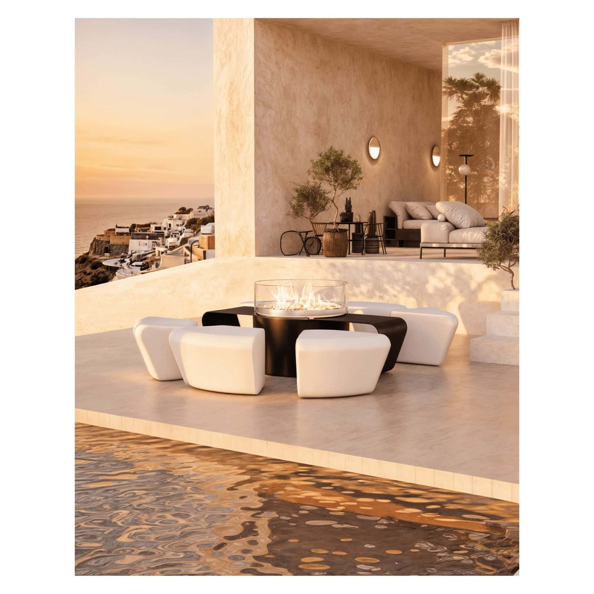 Modern outdoor patio furniture set featuring a round black fire pit table surrounded by six white curved modular poufs, set next to a pool with a sunset view and coastal town in the background.