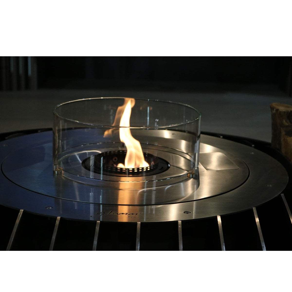 A clear glass wind guard surrounds a small outdoor fire pit with a visible flame, set on a round brushed-metal burner base.
