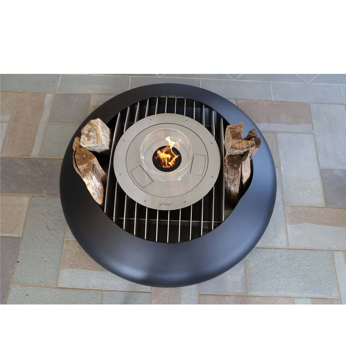 A round outdoor fire pit table with a matte black bowl-shaped outer ring, a metal grate over a small fire in the center, and two logs placed on either side, shown from above on a stone tile patio.