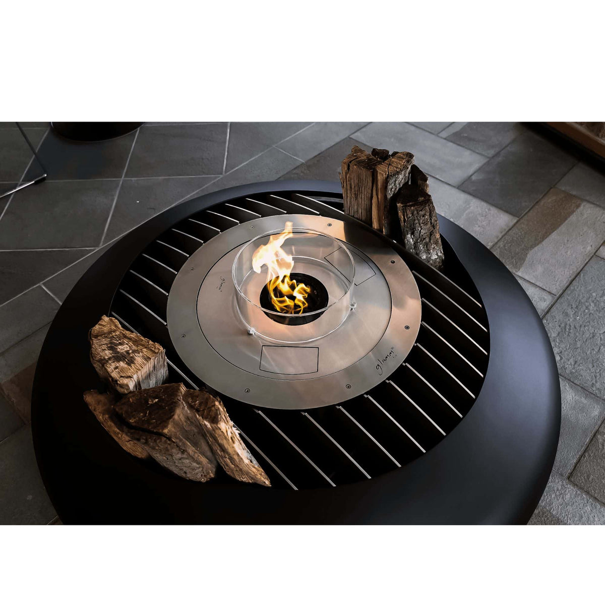 A round black outdoor fire pit table with a glass wind guard and a central flame, shown on a stone patio with logs placed around it.