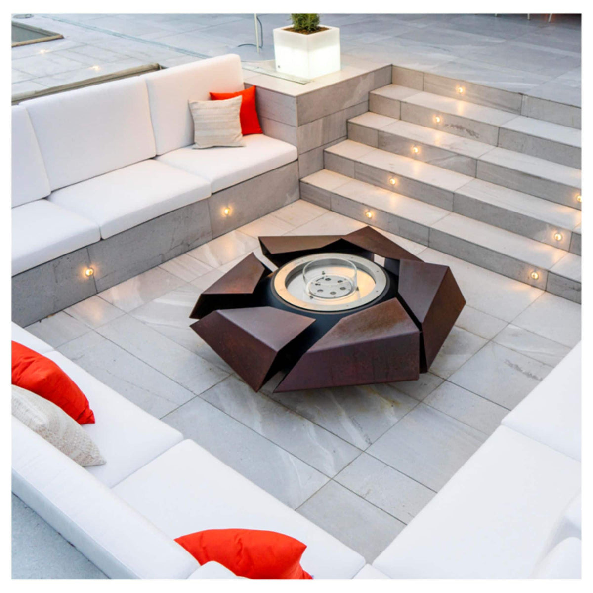A modern outdoor patio scene with a central dark brown geometric fire pit table surrounded by white sectional seating, red and beige accent pillows, and built-in steps with warm lighting.