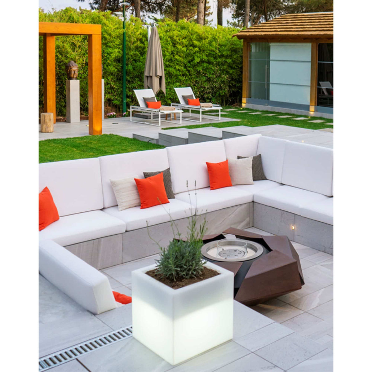 Modern outdoor patio furniture set featuring a white sectional U-shaped sofa with orange and beige accent pillows, a white square illuminated planter with a small plant, a dark brown geometric fire pit table, and a contemporary patio setting with lounge chairs, green hedges, and a wooden structure in the background.