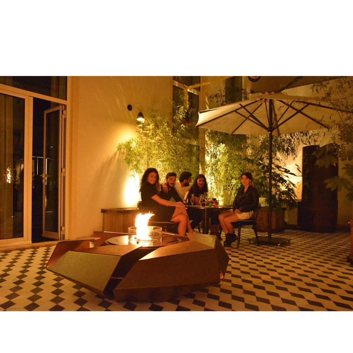 A group of four people sitting and relaxing on a warm, dimly lit patio at night around a modern geometric fire pit table with a flame. The setting features a black and white checkered tile floor, a large patio umbrella, and a warm wall illuminated by plants.