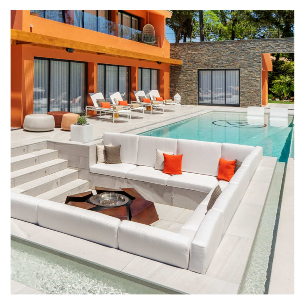 A modern outdoor patio area with a large white sectional sofa, orange accent pillows, a round fire pit table, and a swimming pool beside an orange house.