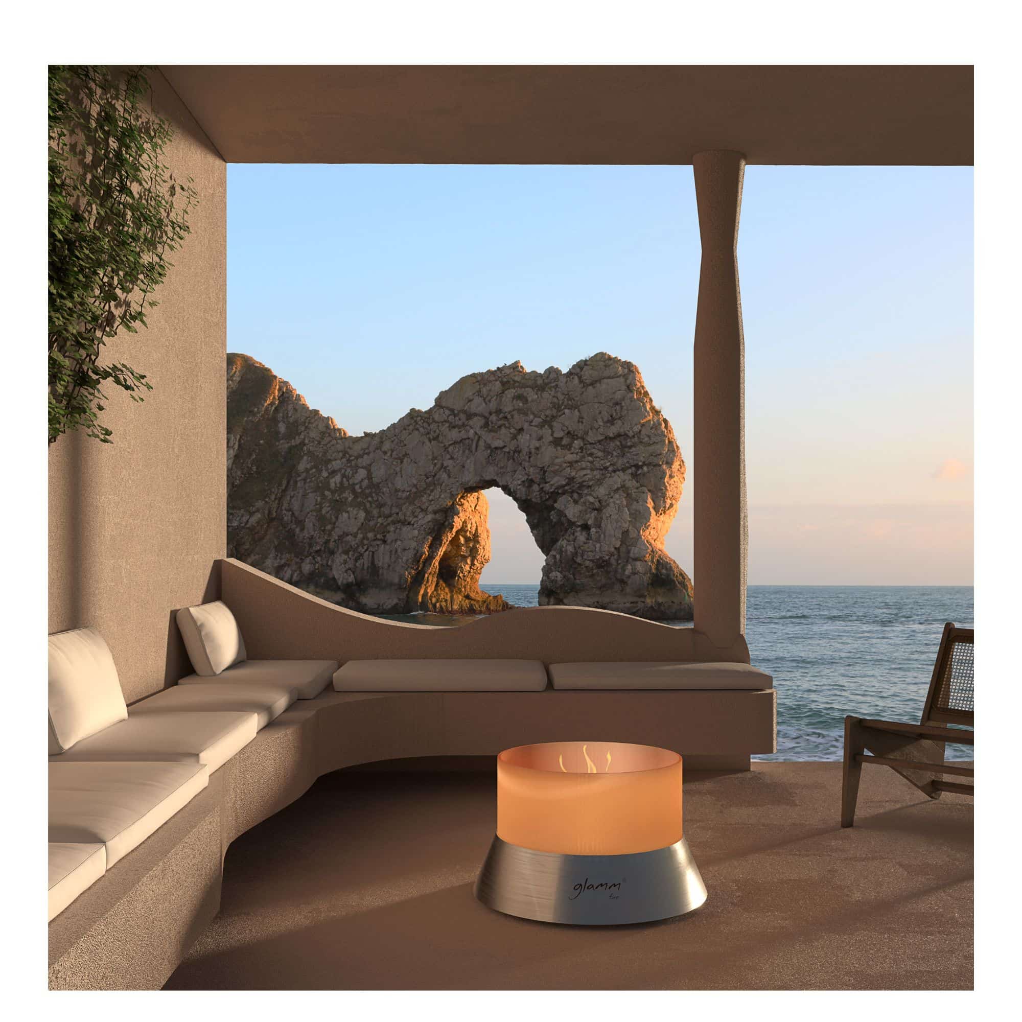 A modern cylindrical firepit sits on a patio overlooking a rocky sea arch and ocean at sunset.