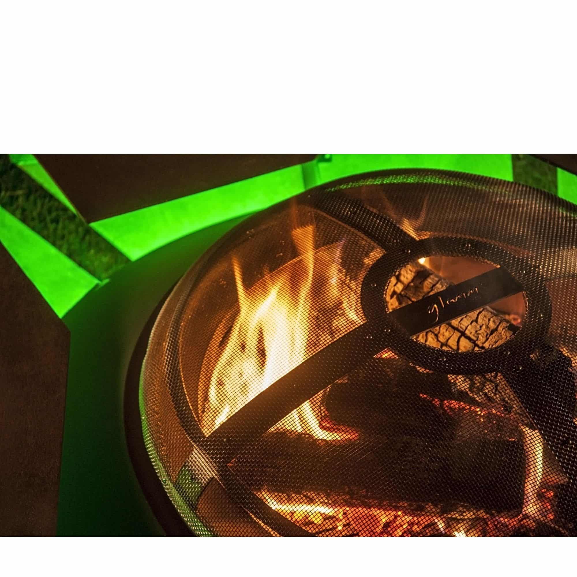 A close-up of a round fire pit with glowing orange flames inside, covered by a black mesh screen. The fire pit is set against a dark background with a bright green light reflection on the left.