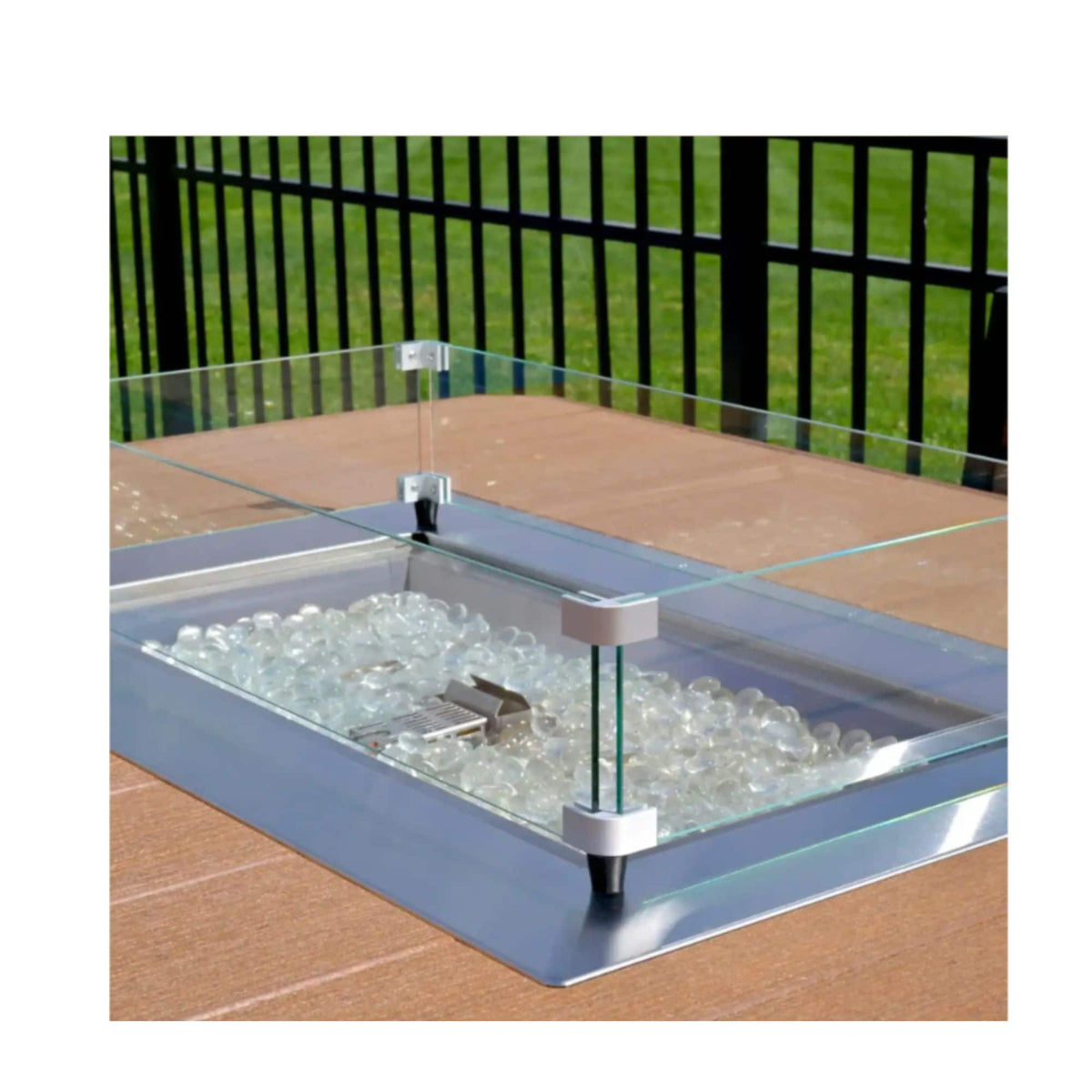 A square glass fire pit insert filled with clear decorative stones, set into a wooden deck with green grass in the background.